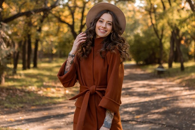 따뜻한 갈색 코트, 가을 유행 패션 거리 스타일을 입고 공원에서 산책하는 곱슬 머리를 가진 아름 다운 세련 된 웃는 마른 여자