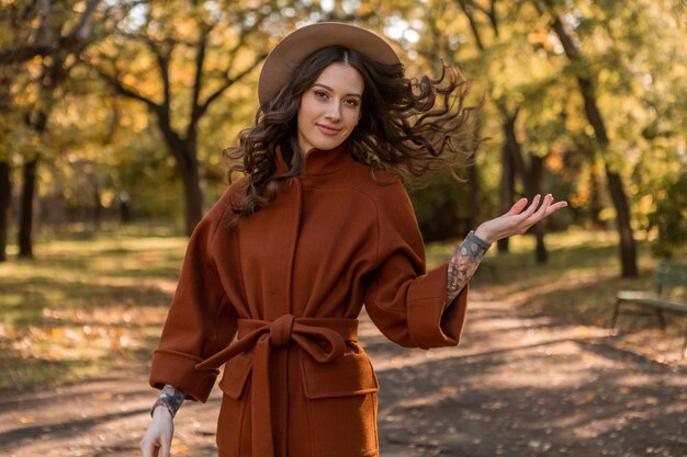 따뜻한 갈색 코트, 가을 유행 패션 거리 스타일을 입고 공원에서 산책하는 곱슬 머리를 가진 아름 다운 세련 된 웃는 마른 여자