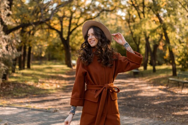 暖かい茶色のコート、秋の流行のファッションストリートスタイルに身を包んだ公園を歩く巻き毛の美しいスタイリッシュな笑顔の細い女性