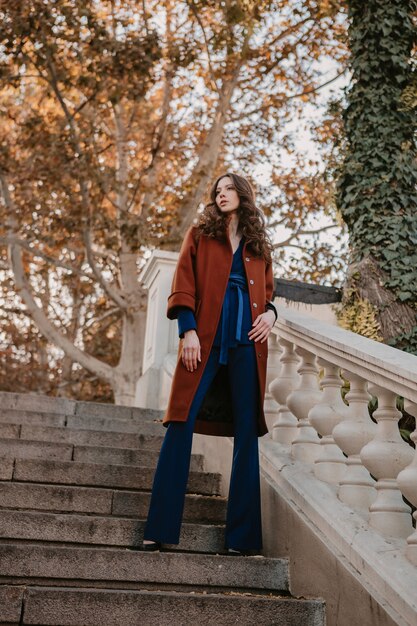 暖かい茶色のコートと青いスーツ、秋の流行のファッションストリートスタイルに身を包んだ通りの階段を歩く巻き毛の美しいスタイリッシュな笑顔の細い女性