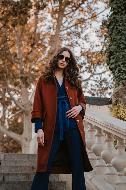 Beautiful stylish smiling skinny woman with curly hair walking in street stairs dressed in warm brown coat and blue suit, autumn trendy fashion street style