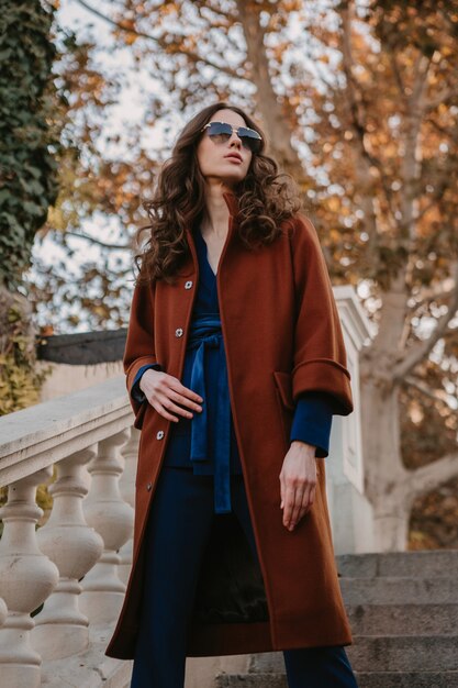 暖かい茶色のコートと青いスーツ、秋の流行のファッションストリートスタイルに身を包んだ通りの階段を歩く巻き毛の美しいスタイリッシュな笑顔の細い女性
