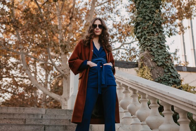 Beautiful stylish smiling skinny woman with curly hair walking in street stairs dressed in warm brown coat and blue suit, autumn trendy fashion street style