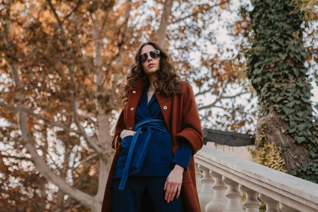 Beautiful stylish smiling skinny woman with curly hair walking in street stairs dressed in warm brown coat and blue suit, autumn trendy fashion street style