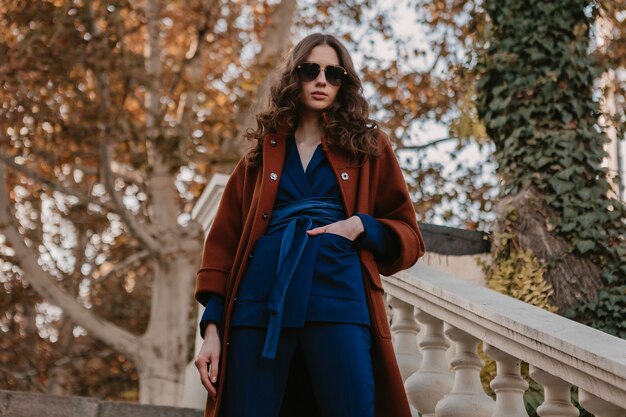 Beautiful stylish smiling skinny woman with curly hair walking in street stairs dressed in warm brown coat and blue suit, autumn trendy fashion street style