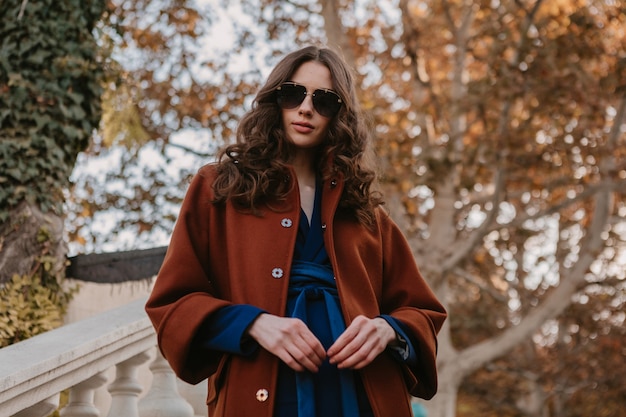 Beautiful stylish smiling skinny woman with curly hair walking in street stairs dressed in warm brown coat and blue suit, autumn trendy fashion street style