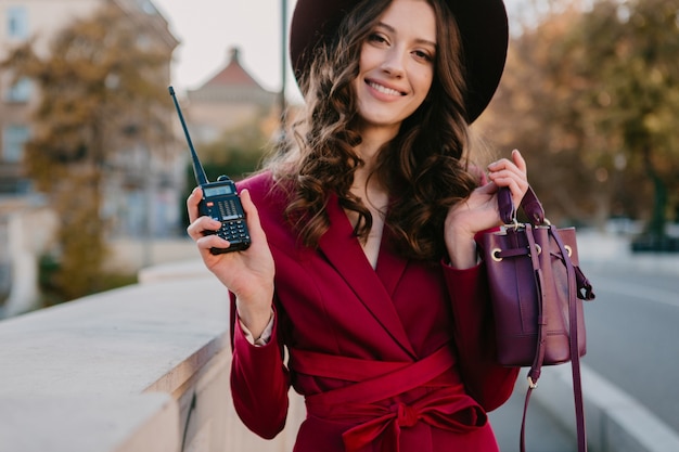 Beautiful stylish woman in purple suit walking in city street, spring summer autumn season fashion trend wearing hat, holding purse holding walkie-talkie