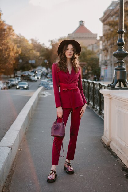 Beautiful stylish woman in purple suit walking in city street, spring summer autumn season fashion trend wearing hat, holding purse