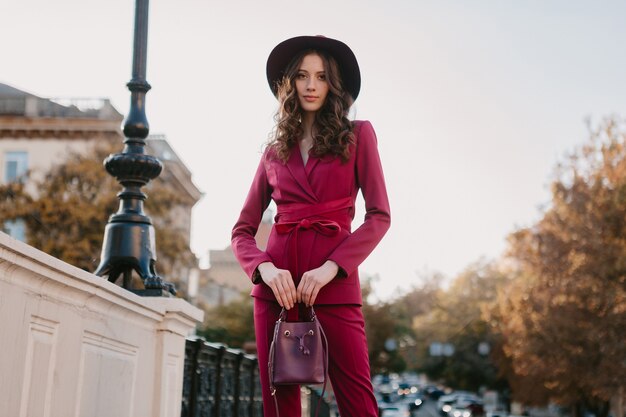 Beautiful stylish woman in purple suit walking in city street, spring summer autumn season fashion trend wearing hat, holding purse