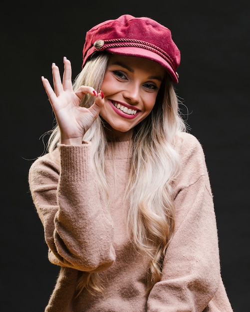 Beautiful stylish young woman wearing pink cap showing ok gesture standing against black background