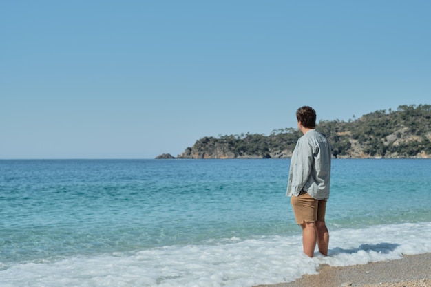 Free photo beautiful summer landscape with a young man standing in coastal waves and looking at the sea against a colorful sky and islands travel background leisure and tourism active lifestyle