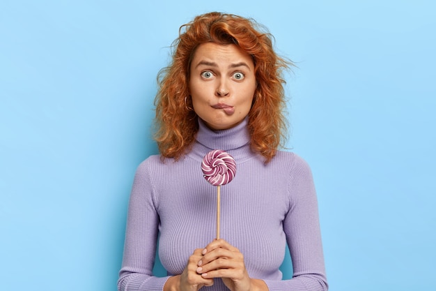 Free photo beautiful surprised female bites lips with appetite, holds purple lollipop on stick, likes sweets