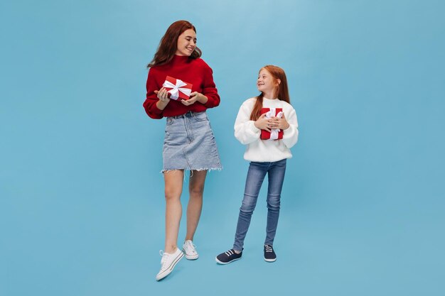 Beautiful tall woman with brunette hair in bright sweater and denim skirt looking at young ginger sister in jeans with gift box