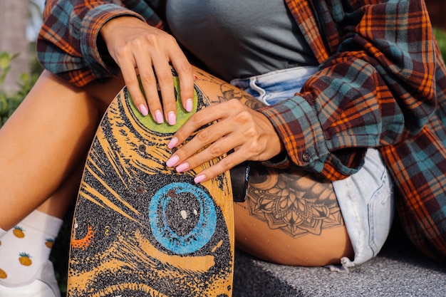 Beautiful tattooed fit woman in jean shorts, plaid shirt sit on stairs at sunset light holding longboard