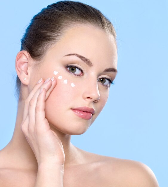 Beautiful teen girl applying cosmetic cream on skin around eyes