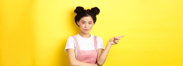 Beautiful teenage asian girl with bright makeup pointing finger left at banner and smiling yellow ba