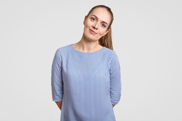 Beautiful teenage girl has dark hair combed in pony tail, wears striped blue shirt, keeps hands behind back