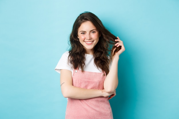 Beautiful teenage girl waiting for summer, playing with curly hair and smiling white teeth, having cute blush and glam make up on face, standing against blue background