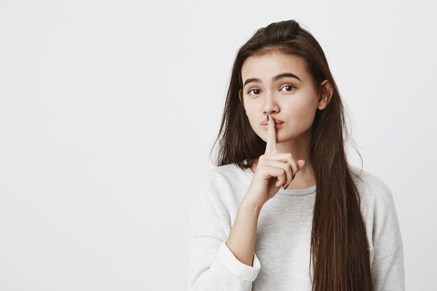 Beautiful teenage woman with dark long hair dressed casually holding index finger on lips, asking to keep silence, not tell anyone her secret, raising brows, saying Shh. Confidential information