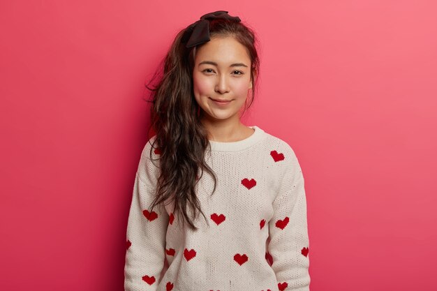 Beautiful tender Asian girl with long pony tail, rouge cheeks, wears comfortable jumper with hearts, stands against pink background