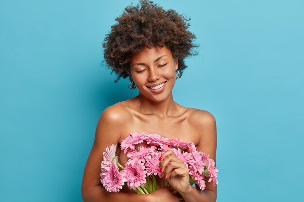 Beautiful tender woman stands with closed eyes, naked body, carries pink gerbera daisy flowers, smiles happily, has healthy dark skin, enjoys pleasant fragrancy, 