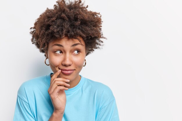 Beautiful thoughtful young African American woman looks aside with pensive dreamy expression has curly hair dressed casually considers something isolated over white wall blank copy space