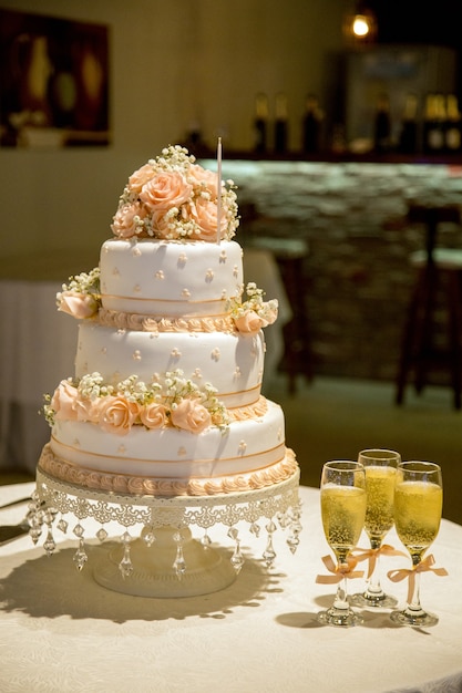 A beautiful three-layered cake with rose decorations and glasses of champagne on the table