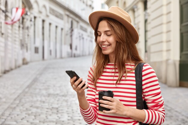 Beautiful tourist strolls on city streets, uses online navigator for finding right way, holds cell phone