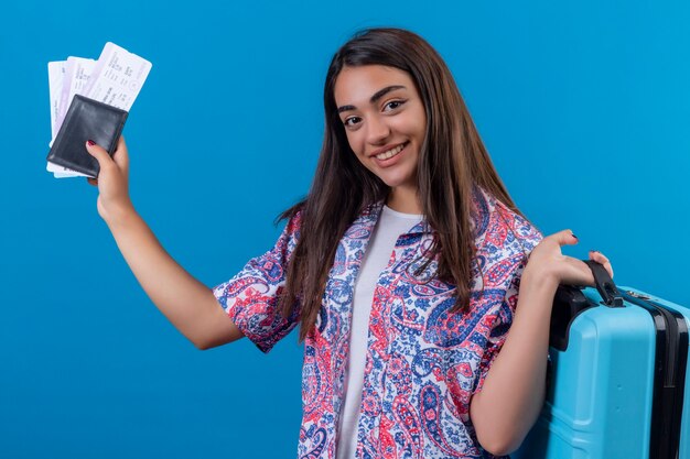 Beautiful tourist woman holding travel suitcase and passport with tickets  with smile on face happy and positive travel concept standing over blue space