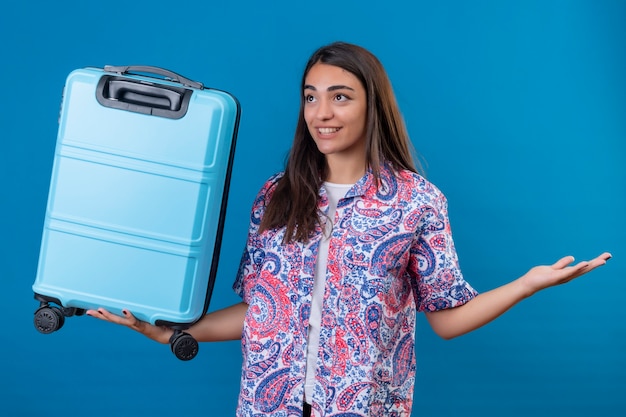 孤立した青い空間の上に元気に立って笑顔の側面に彼女の手を広げて旅行スーツケースを保持している美しい観光女性