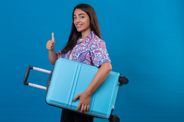 Beautiful tourist woman standing with travel suitcase smiling cheerfully showing thumbs up  over isolated blue space