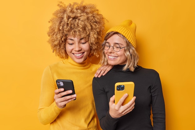 Beautiful two female friends use mobile phones browse internet type sms message dressed in casual turtlenecks smile gladfully isolated over vivid yellow background watch video via media app