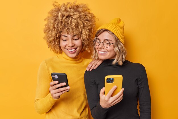 Beautiful two female friends use mobile phones browse internet type sms message dressed in casual turtlenecks smile gladfully isolated over vivid yellow background watch video via media app