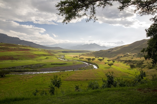 Beautiful view of the river, trees, and grasses surrounded by mountains with a cloudy blue sky