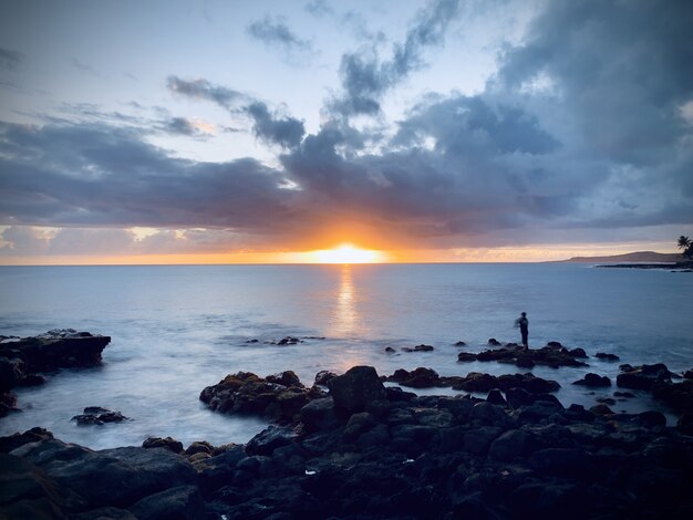 Beautiful view of the sunset in the cloudy sky over the calm ocean by the rocky shore