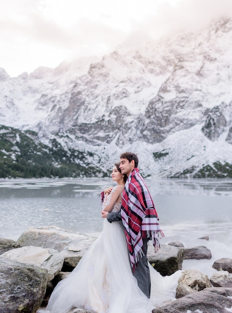 Beautiful wedding couple covered with bright blanket is standing in front of frozen lake surrounded with snowy mountains