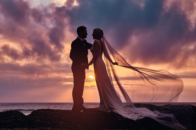 Beautiful wedding couple holds each other hands standing on the beach against the amazing sunset.