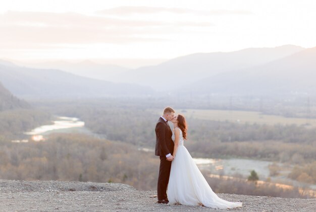 Beautiful wedding couple is kisssing on the hill with a view of a picturesque landscape in the dusk