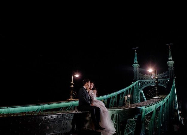 Beautiful wedding couple is sitting on the illuminated bridge on the dark night and kissing