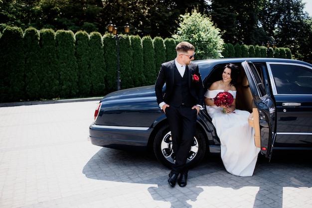 赤い花束とエレガントな結婚式の服を着て、晴れた日に美しい結婚式のカップルが黒い車で笑っています。