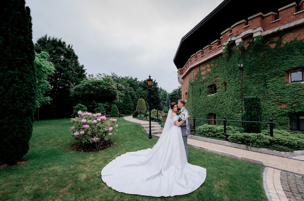 Beautiful wedding couple is standing in the green park near the building fully covered with leaves