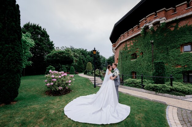 Beautiful wedding couple is standing in the green park near the building fully covered with leaves