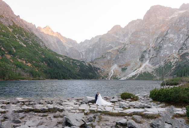 高山の湖の美しい景色の前に美しい結婚式のカップルが立っています。