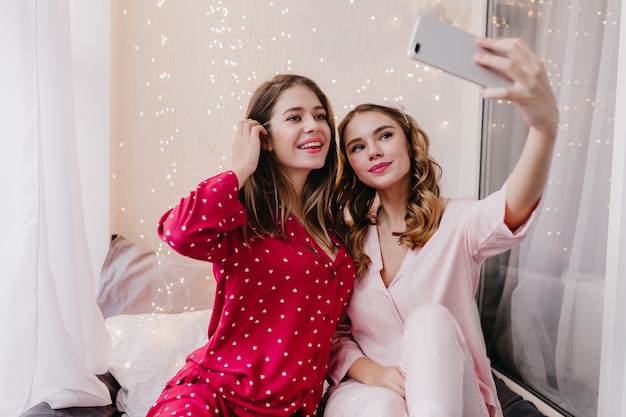 Beautiful white girl posing with pleasure early in morning in her room. Cute curly woman using phone for selfie with long-haired sister.