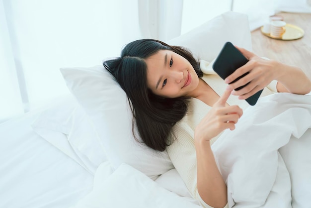 아름다운 흰색 셔츠 아시아 여성은 흰색 침대 휴가 생활 방식으로 스마트폰을 잡고 대화를 즐깁니다.