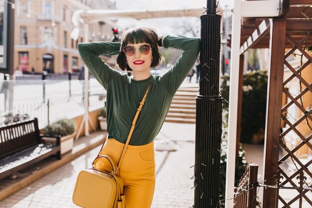 通りに手を上げてポーズをとって興味を表す美しい白人女性。黄色のハンドバッグを保持している緑のセーターの魅力的な黒髪の女の子の屋外写真。