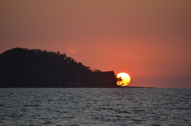 Beautiful wide silhouette shot of an islet covered with trees on by the sea under sky during sunset