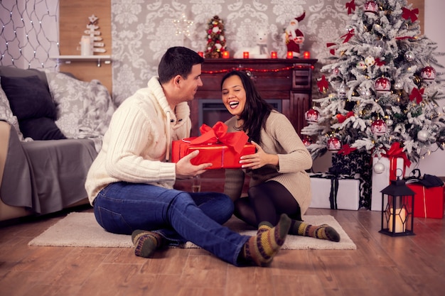 Beautiful wife laughing and giving her husband christmas gift box. Christmas decoration.