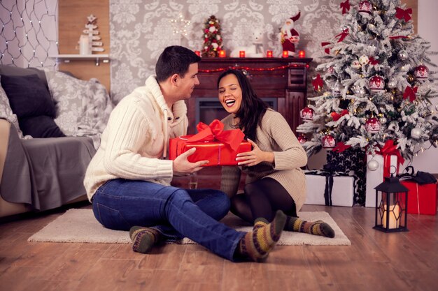 Beautiful wife laughing and giving her husband christmas gift box. Christmas decoration.