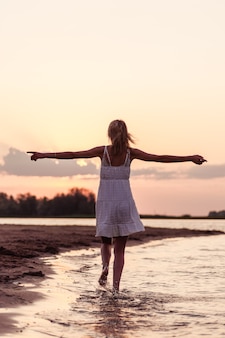 Beautiful woman on the beach rear view. a young slender young blonde in a white dress runs along the river bank against the background of the sunset and with her hands raised.
